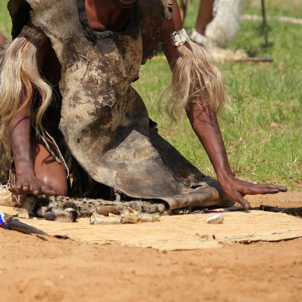 A traditional African diviner dressed in animal hides casts bones onto a mat during a ritual divination practice outdoors.
