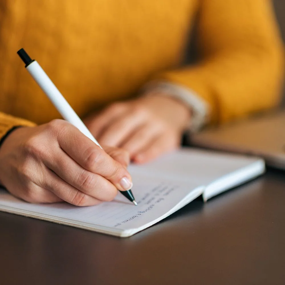 A person in a yellow sweater writing with a black and white pen in a notebook, representing journaling or automatic writing as a form of divination or self-reflection.