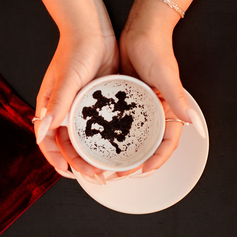 A close-up of hands holding a white teacup with distinct coffee ground patterns inside, used for tasseography or coffee ground reading.