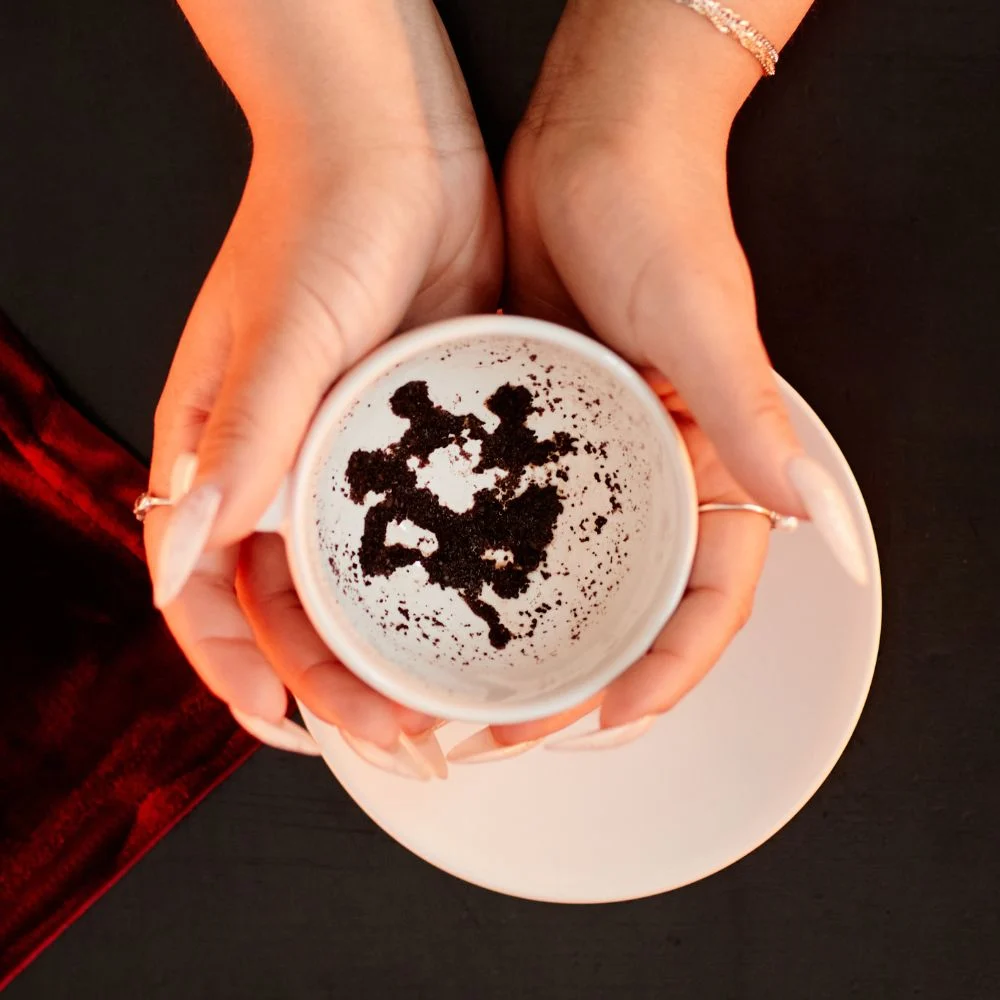 A close-up of hands holding a white teacup with distinct coffee ground patterns inside, used for tasseography or coffee ground reading.