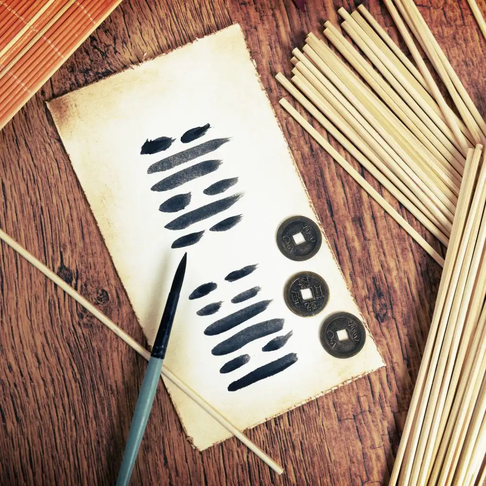 A traditional I Ching divination setup with black brush-painted hexagrams on aged paper, surrounded by bamboo sticks and three Chinese coins on a wooden table.