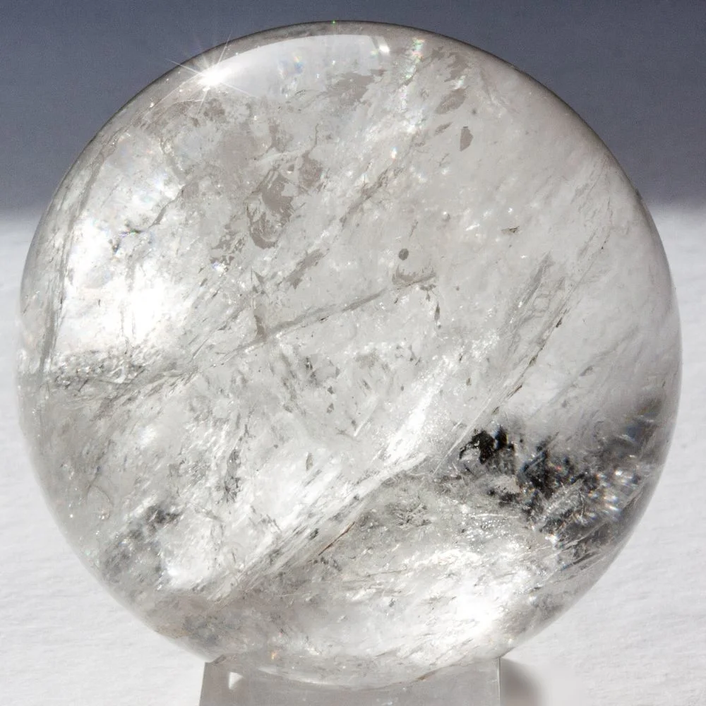 A clear quartz crystal ball with natural inclusions and light reflections, placed on a white surface and used for scrying or divination.