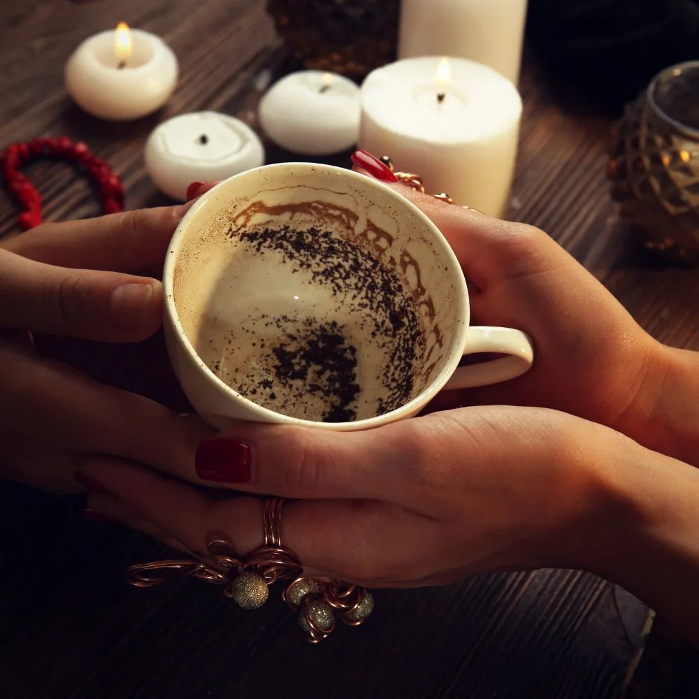A close-up of two hands holding a teacup with leftover coffee grounds inside, used for tasseography, with lit candles and beads in the background.