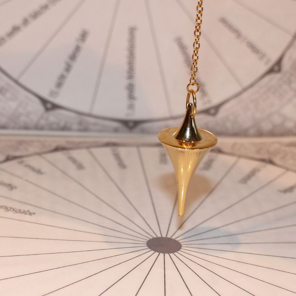 A gold divination pendulum suspended over a printed dowsing chart, used for answering yes/no questions or locating objects.