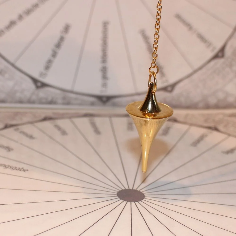 A gold divination pendulum suspended over a printed dowsing chart, used for answering yes/no questions or locating objects.
