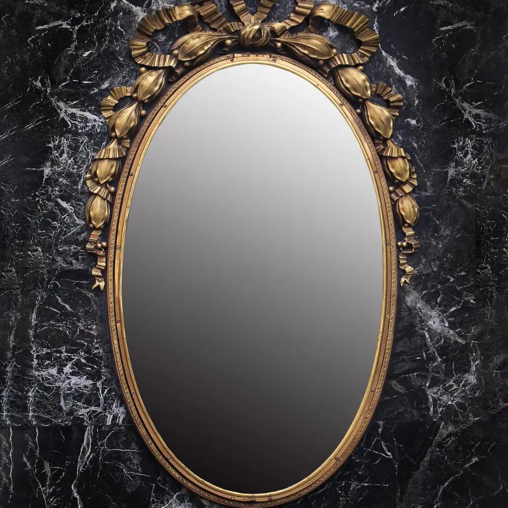 An ornate oval mirror with a gold floral frame mounted on a black marble wall, often used in scrying or mirror gazing practices.