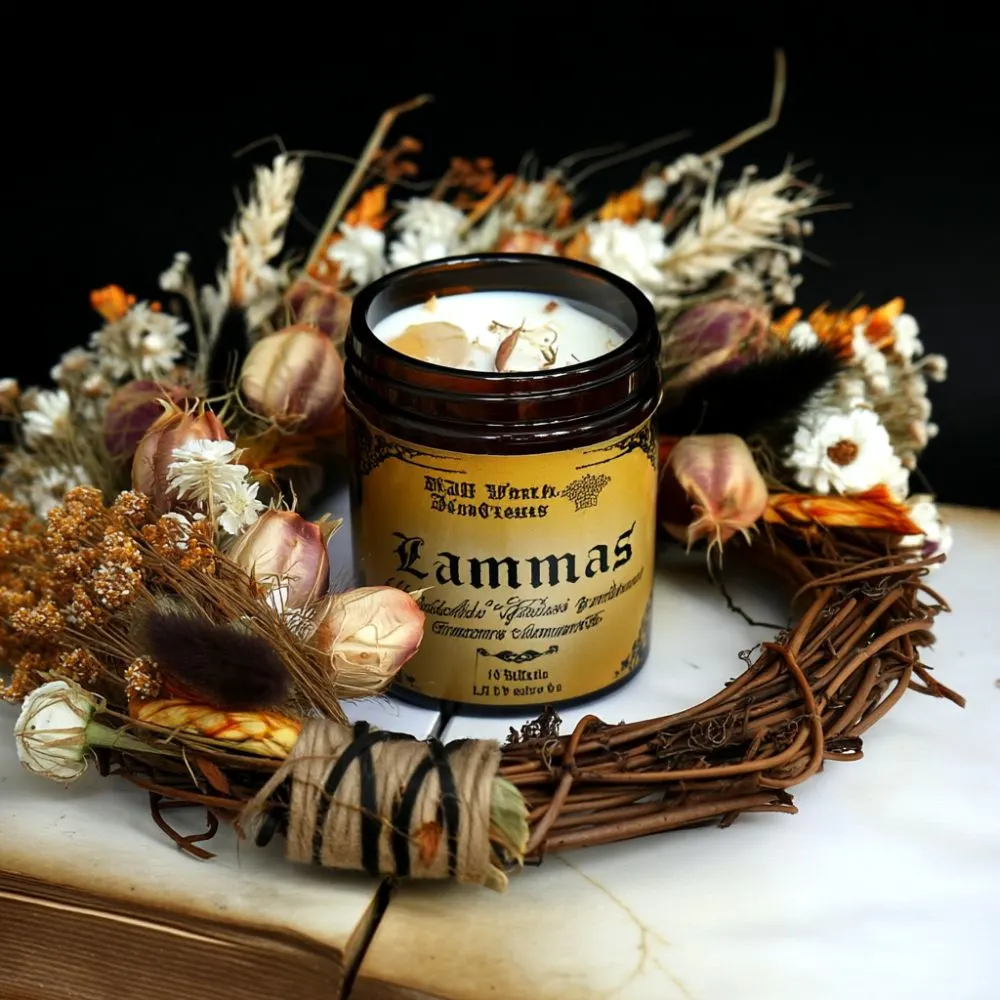 Dark amber glass candle labeled 'Lammas' surrounded by dried flowers, herbs, and a rustic grapevine wreath. The candle is decorated with botanical elements and sits on a marble surface, creating a warm and magical harvest atmosphere.