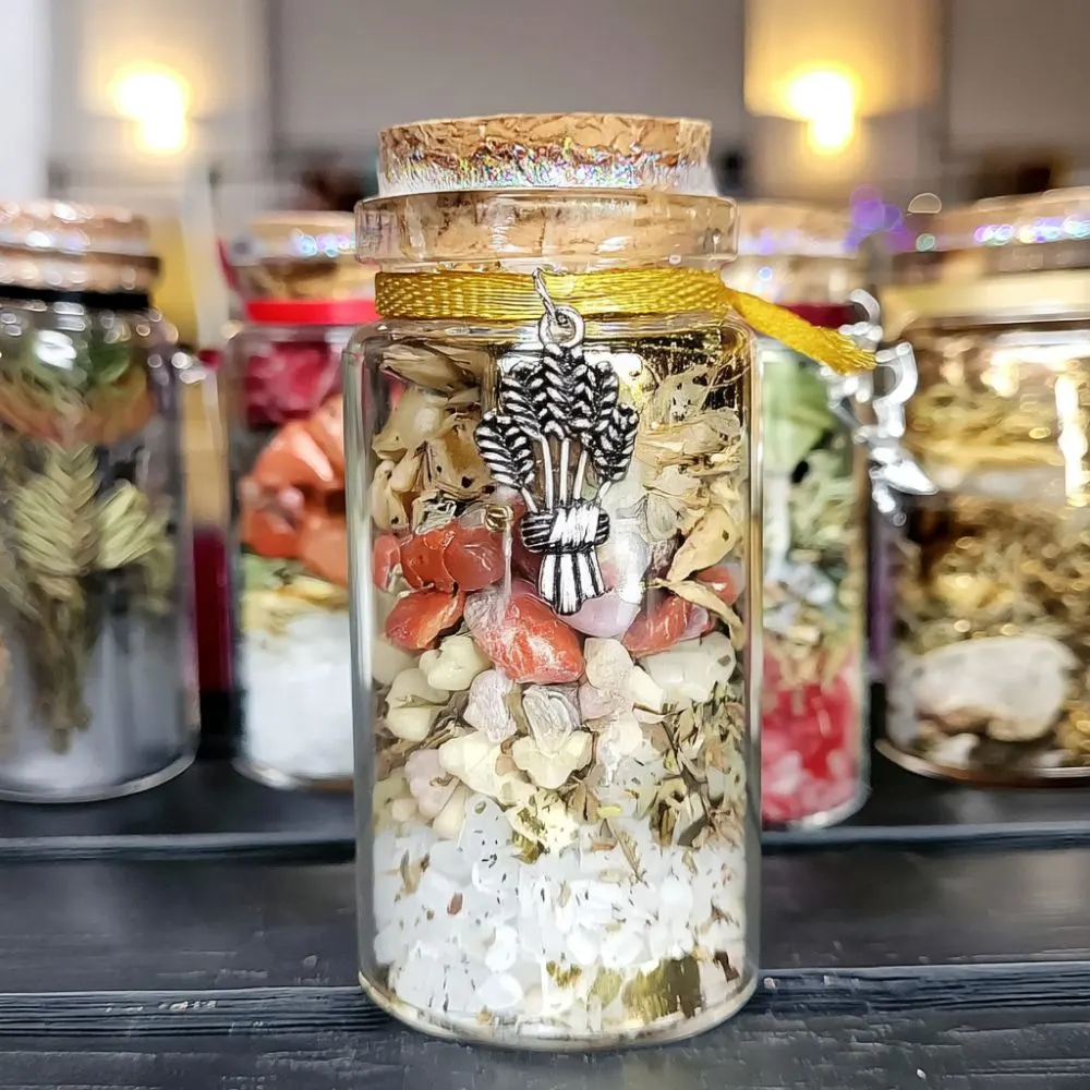 Glass spell jar filled with layered herbs, crystals, and salts, sealed with a cork lid and wrapped with golden string. A silver wheat charm hangs on the front, symbolizing Lammas. Blurred jars of similar contents are in the background, softly lit.